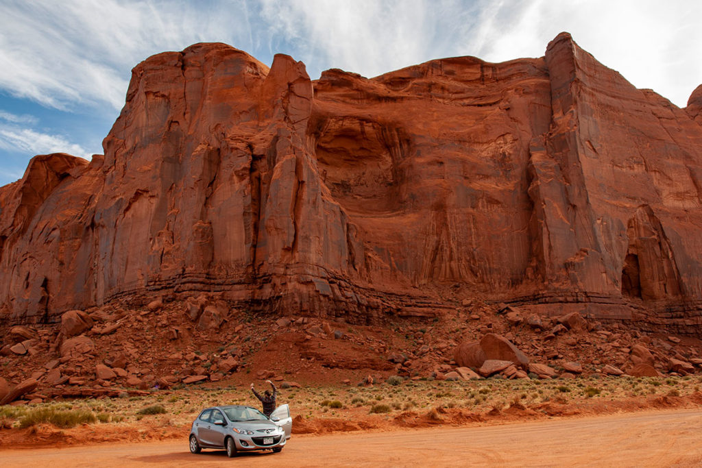 man stretching next to silver sedan at the base of a red-orange cliff
