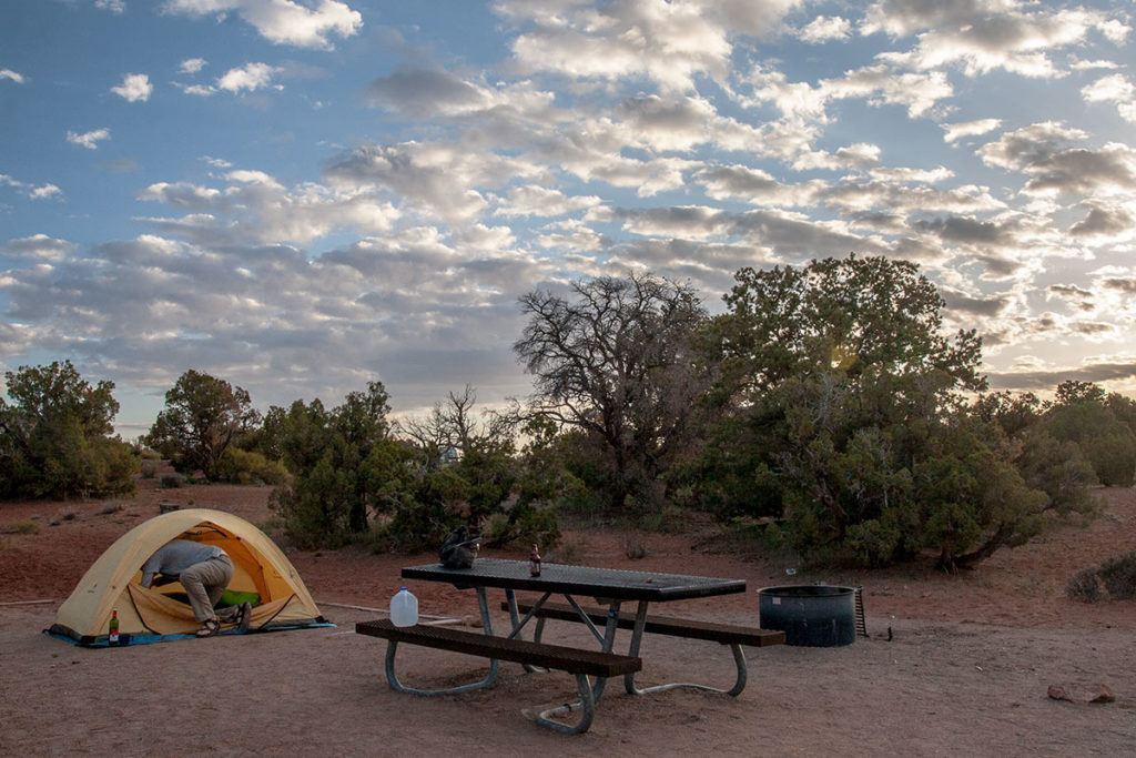 the day's last light lines a sky of clouds in bright white above a campsite surrounded by stubby pinon pines, a yellow backpacking tent set up beside a picnic table bare except for a gallon jug of water and a beer
