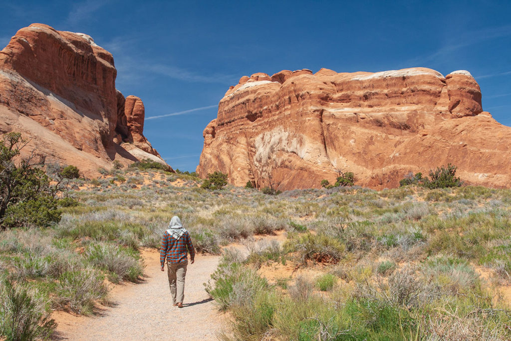 a man in long-sleeved shirt and bare feet walks a gravel trail through scrubland towards two massive orange rock outcroppings, weathered and textured with white highlights and shadowed crevices