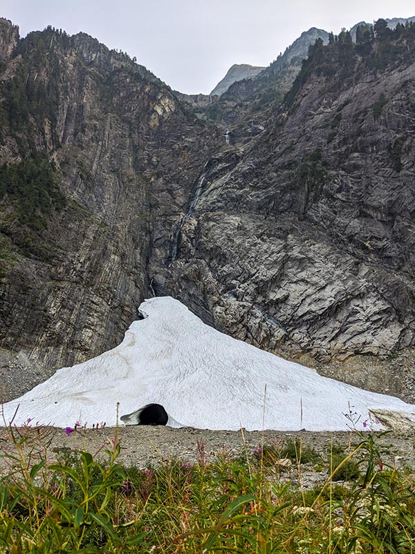 a triangular snowfield wedged against a rocky cliff opens a round mouth created by a stream running beneath it, tiny people beside the ice cave's opening