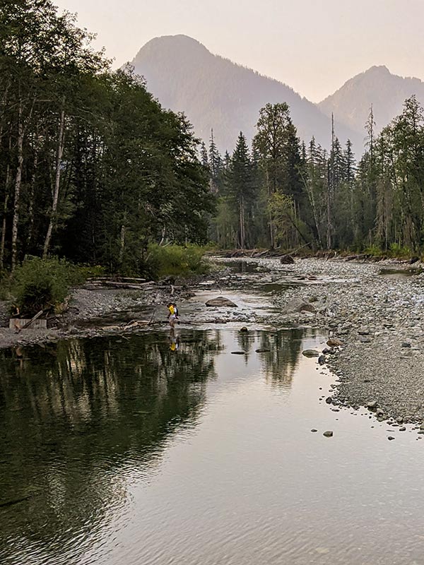 man in yellow shirt carries a child across a placid shin-deep river below a mountain vista tinted pink by smoke